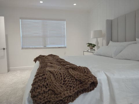 White faux-wood-blinds in a minimalist bedroom. The bed had white sheets, with a brown knitted cover